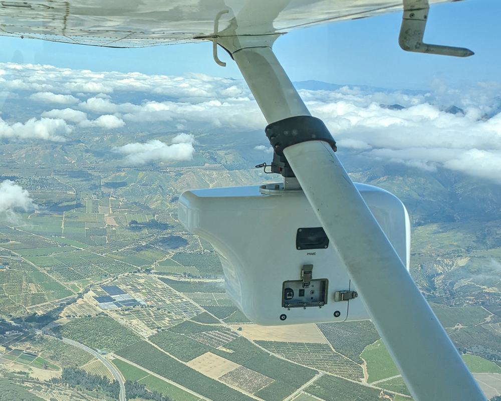 View from beneath a plane with a surveying device attached underneath, flying high above a vast landscape.
