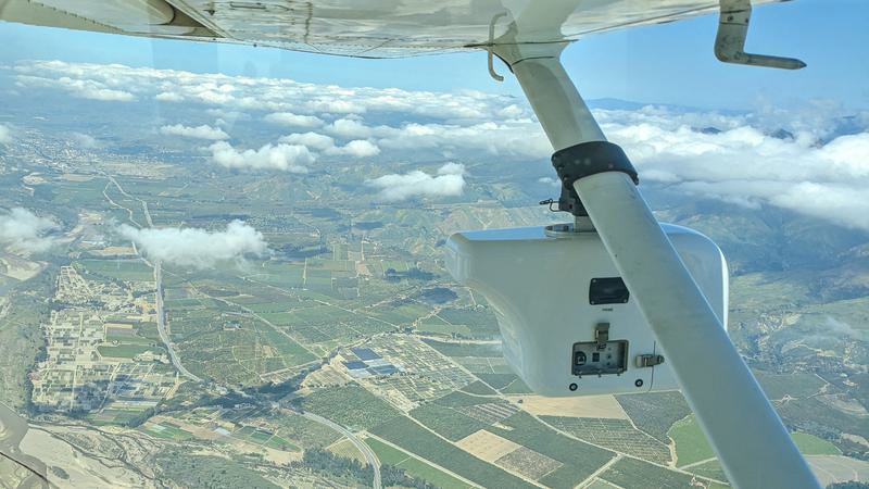 View from beneath a plane with a surveying device attached underneath, flying high above a vast landscape.