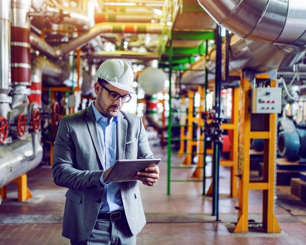 Man in a hard hat and business casual attire looking at a tablet inside an oil refinery