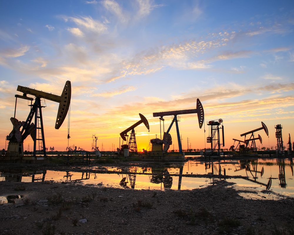 Silhouettes of five oil well pump jacks and various oil drilling equipment against a sunset background