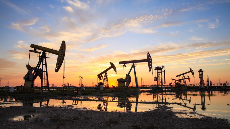 Silhouettes of five oil well pump jacks and various oil drilling equipment against a sunset background