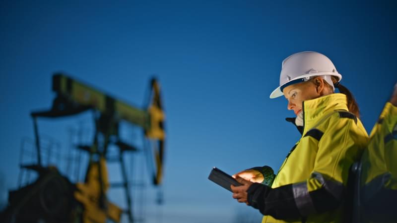 A woman in a hard hat and work jacket looks at an ipad with an oil well in the background