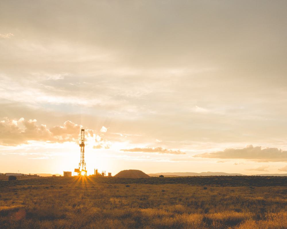 An oil well stands several hundred yards in the distance across a grassy plain with the sun setting in the background