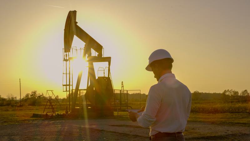 A person in a hard hat looking down at a tablet in front of an oil rig backlit by a warm, bright sunrise.